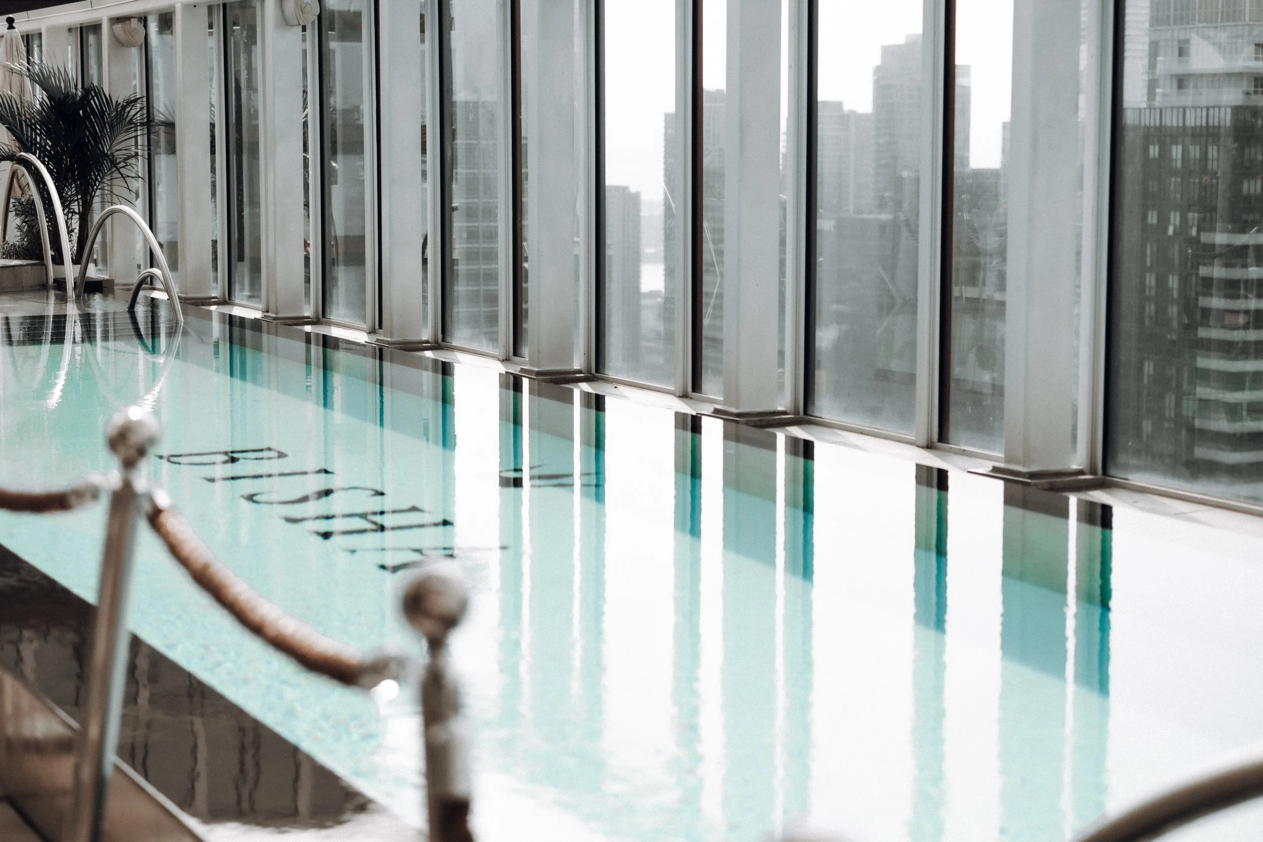 Indoor hotel or apartment pool with metal handrails, a row of large windows showing city buildings outside, and a pool rope barrier in the foreground.