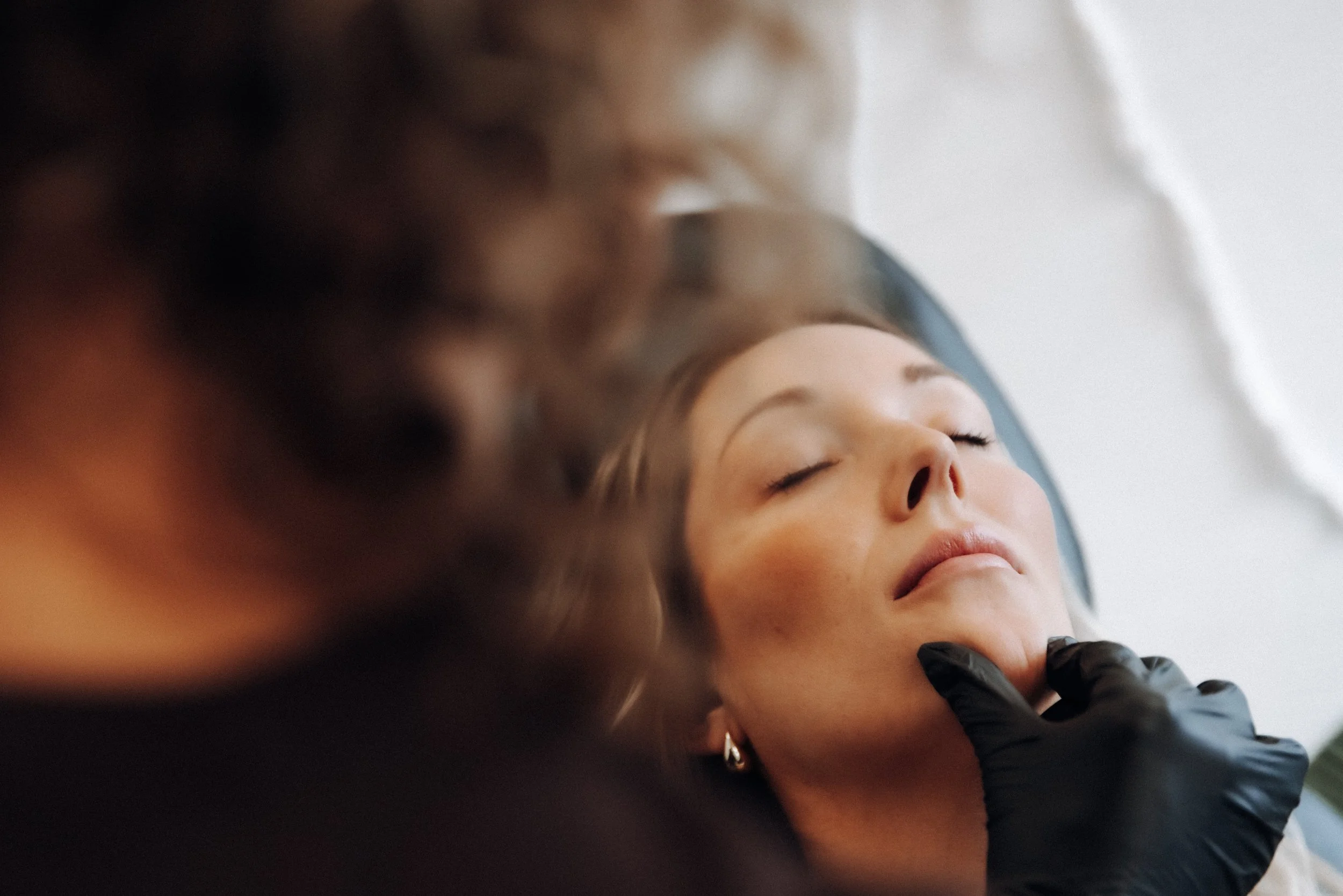A woman with closed eyes receives a facial treatment from a person wearing black gloves. The woman appears relaxed with a calm expression.