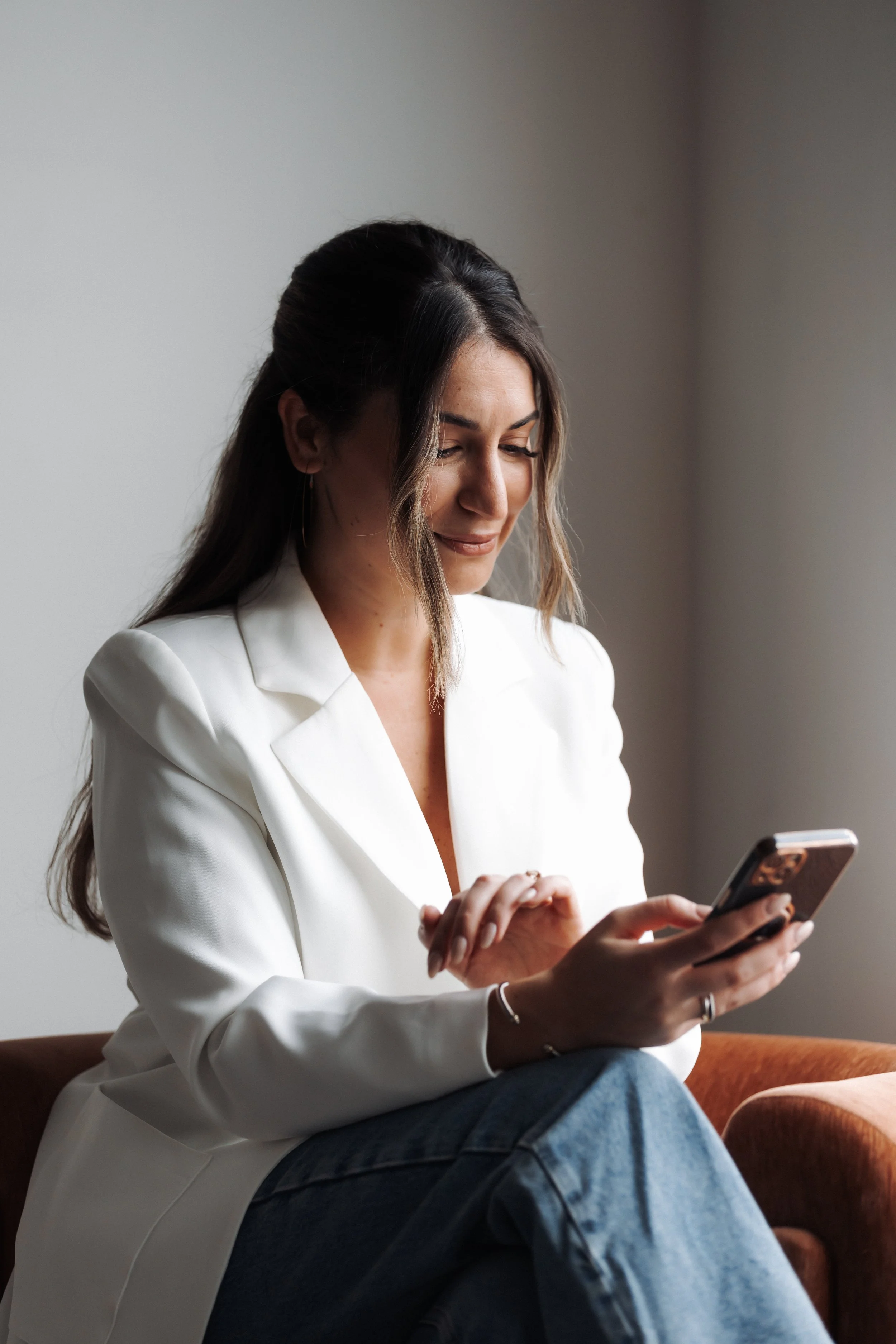 A woman with long, dark hair wearing a white blazer and jeans sitting on a brown chair, looking at her smartphone and smiling.