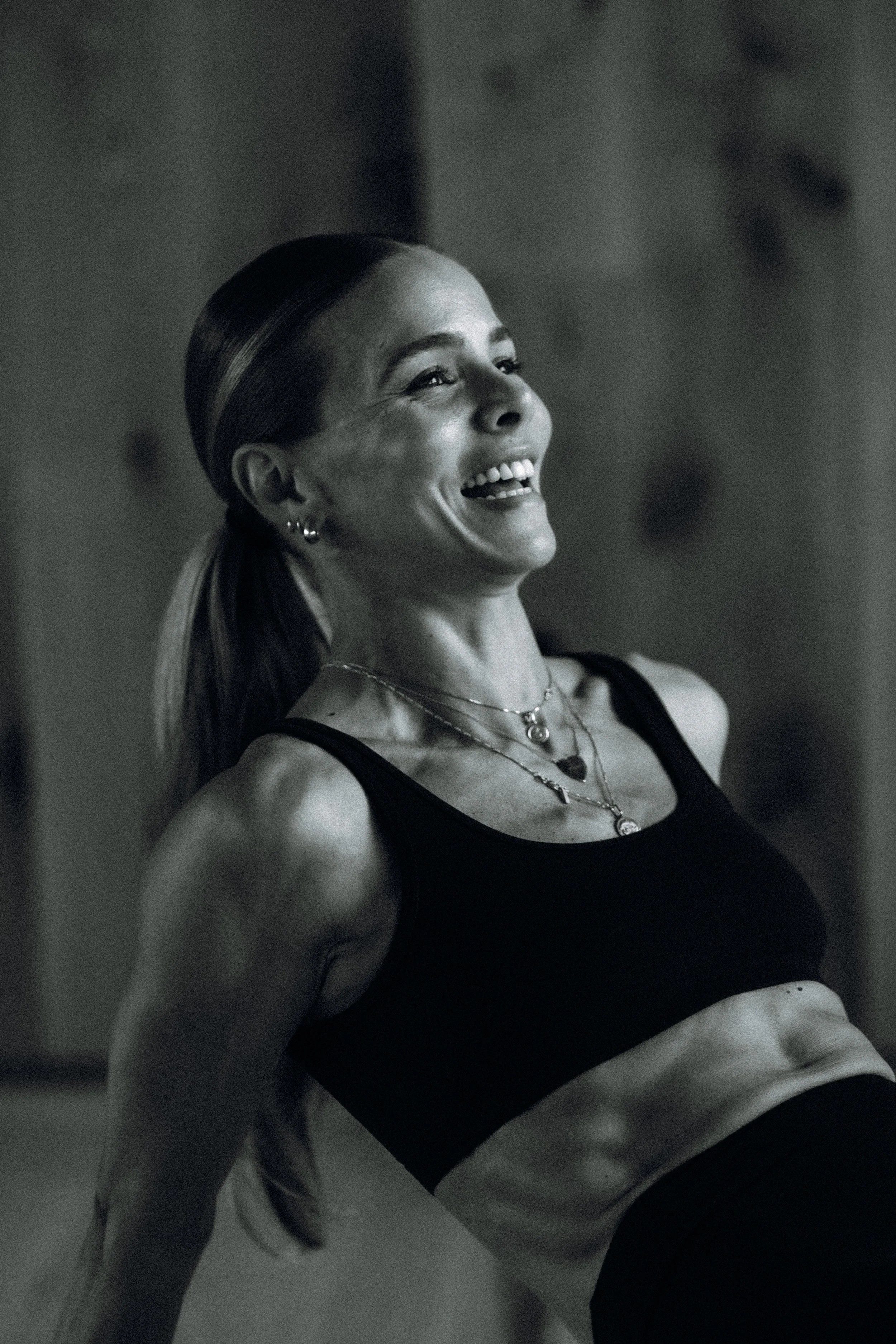 A woman with her hair in a ponytail, smiling and looking upwards, wearing a black sports bra and layered necklaces, in what seems to be a gym or workout space.