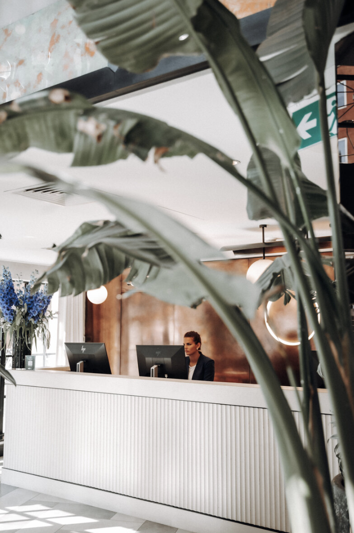 A woman working at a reception desk behind large green indoor plants with brown and white walls in the background.