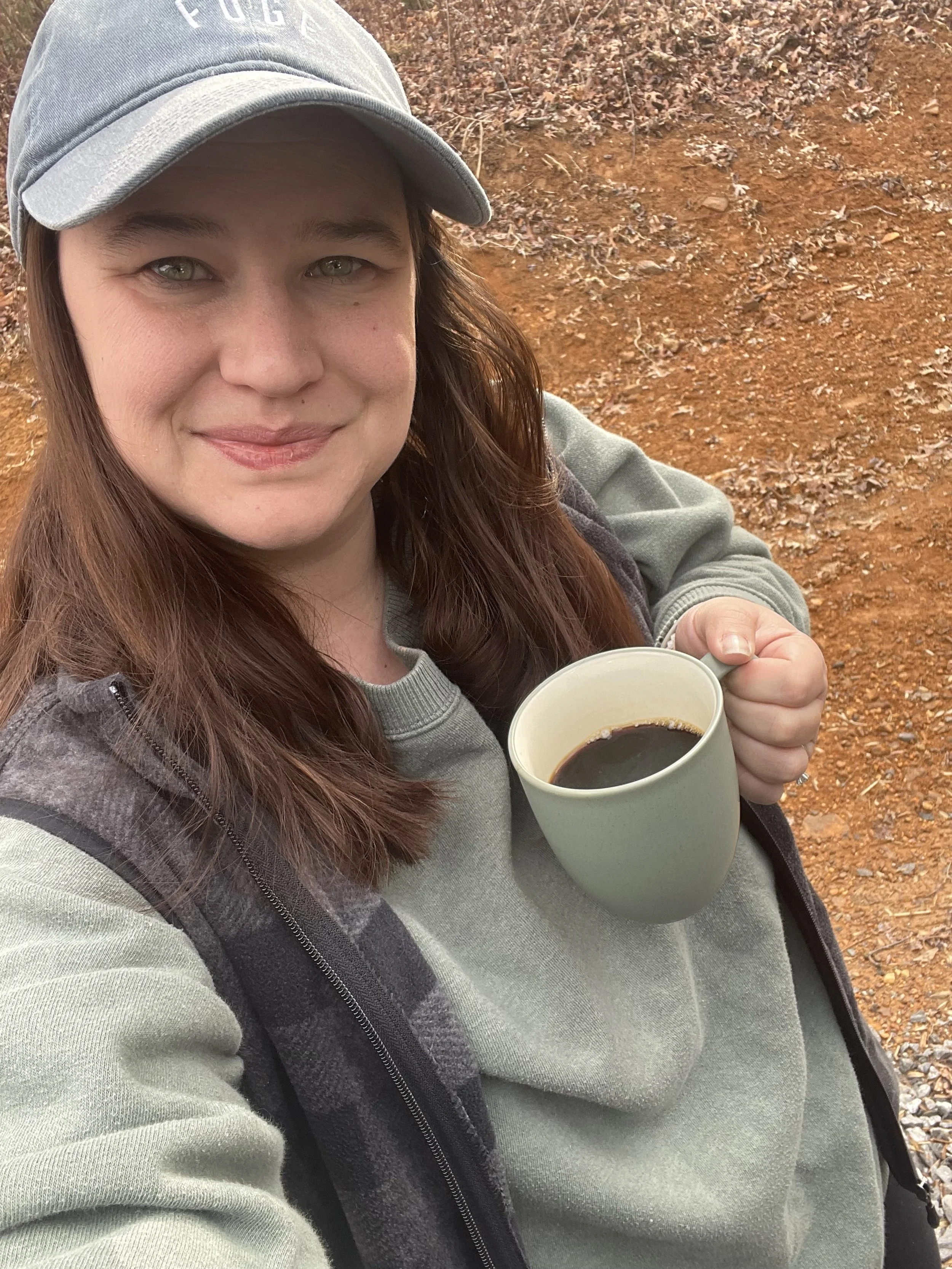 A woman with long brown hair wearing a gray baseball cap and a gray sweatshirt, holding a mug of black coffee, standing outdoors on rocky red earth ground.