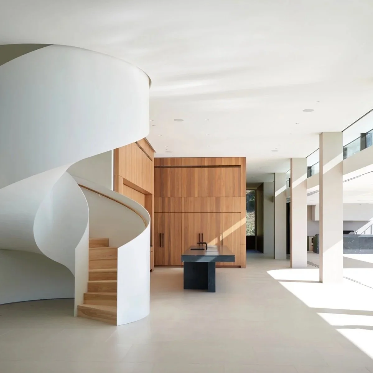 Modern interior with white spiral staircase, wooden cabinets, black table with faucet, and large windows letting in natural light.