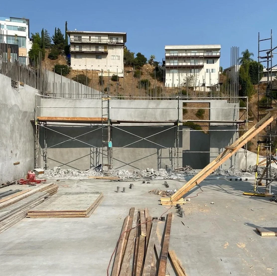 Construction site with scaffolding and building materials, overlooking a hillside with residential buildings under clear sky.