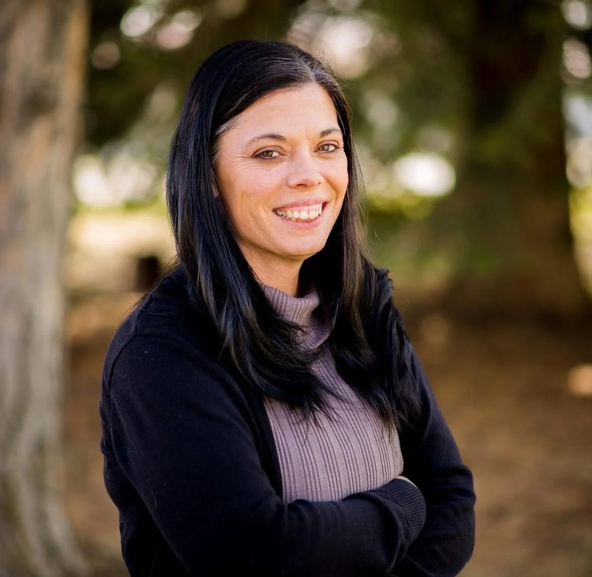 A woman with long black hair smiling outdoors with trees in the background.