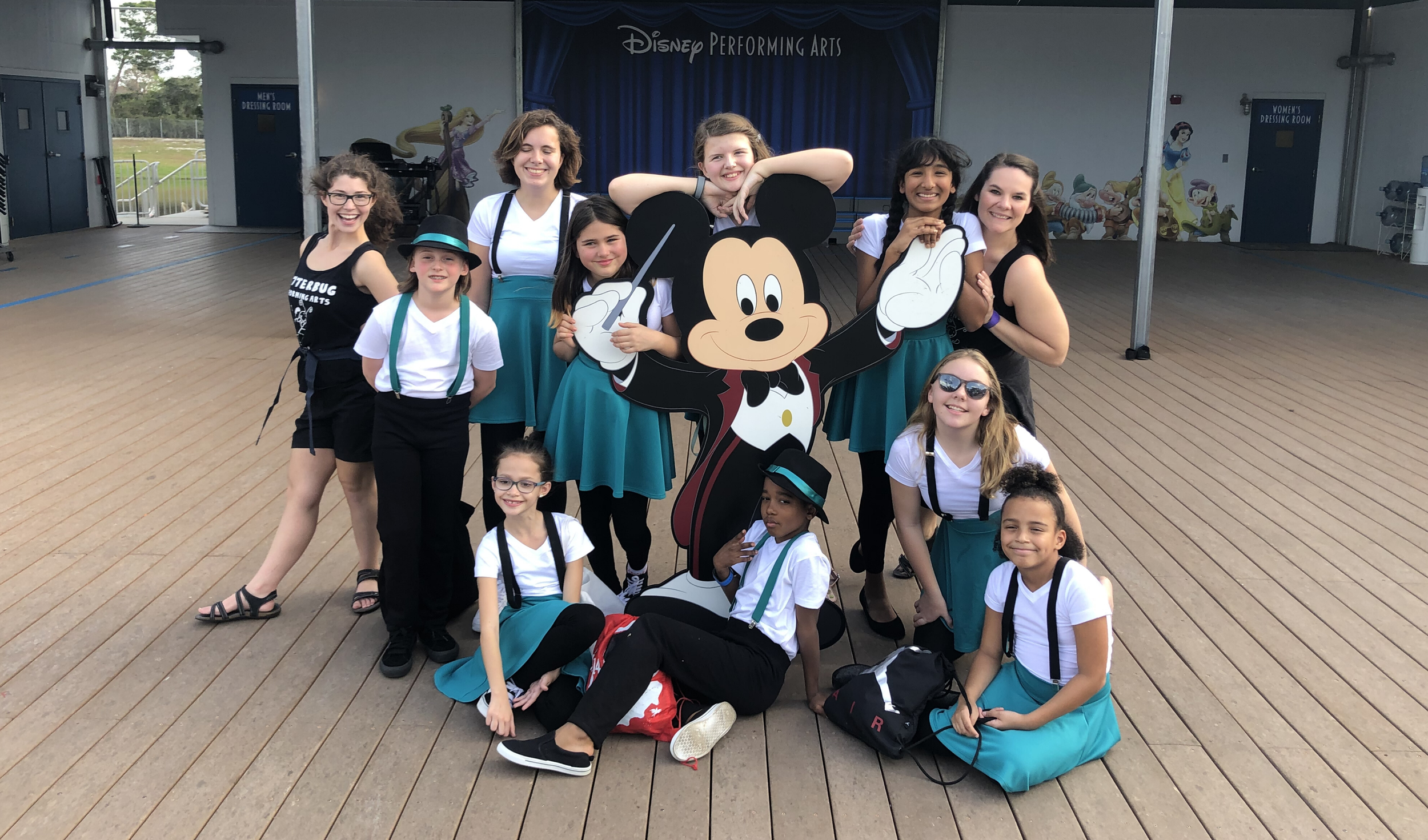 Group of young girls posing around a Mickey Mouse cutout on a wooden outdoor stage at Disney Performing Arts, with Disney artwork on the walls in the background.