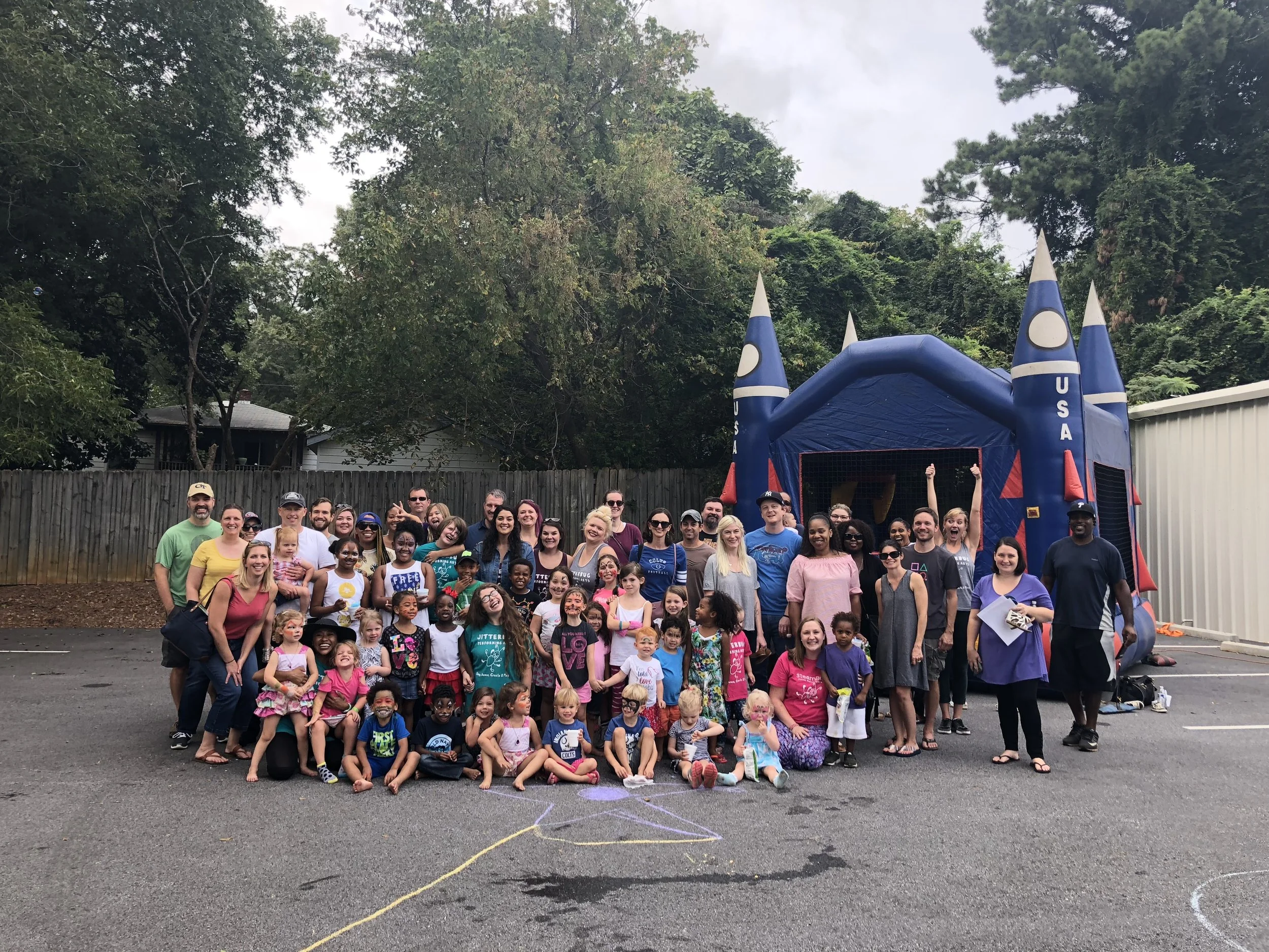 A large group of children, teenagers, and adults gathered outdoors on a paved area in front of a blue inflatable bounce house with a space theme, surrounded by trees and a wooden fence.