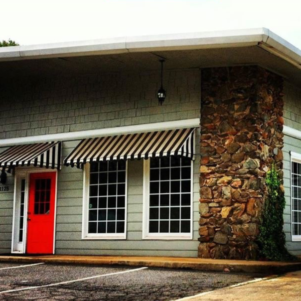Exterior view of a building with a stone column, beige siding, a red door, two large windows, and a black and white striped awning.