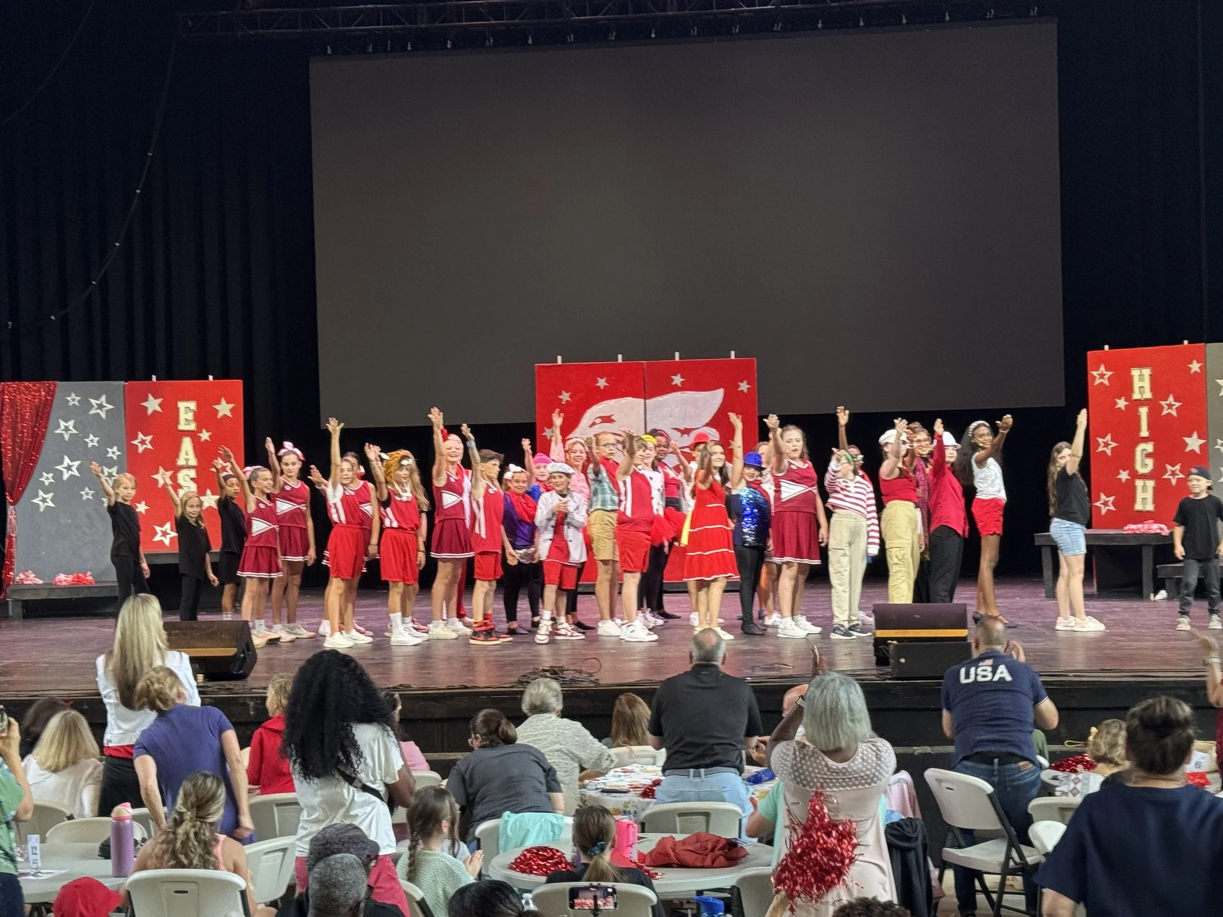 Children's choir performing on stage during a holiday show, dressed in red, black, and festive attire, with Christmas decorations and an audience watching.