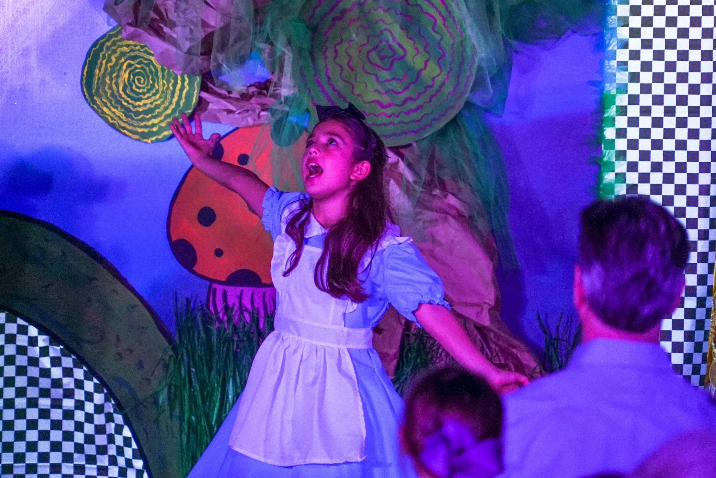 A young girl with long dark hair, wearing a light blue dress, appears to be singing or speaking expressively with her arms raised. She stands in front of a colorful, whimsical background resembling a garden with large cartoonish flowers and a ladybug, under vibrant lighting. Two people are partially visible in the foreground, facing her.