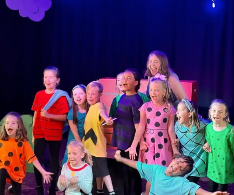Children and a woman laughing and singing on a stage, dressed in colorful costumes for a performance.