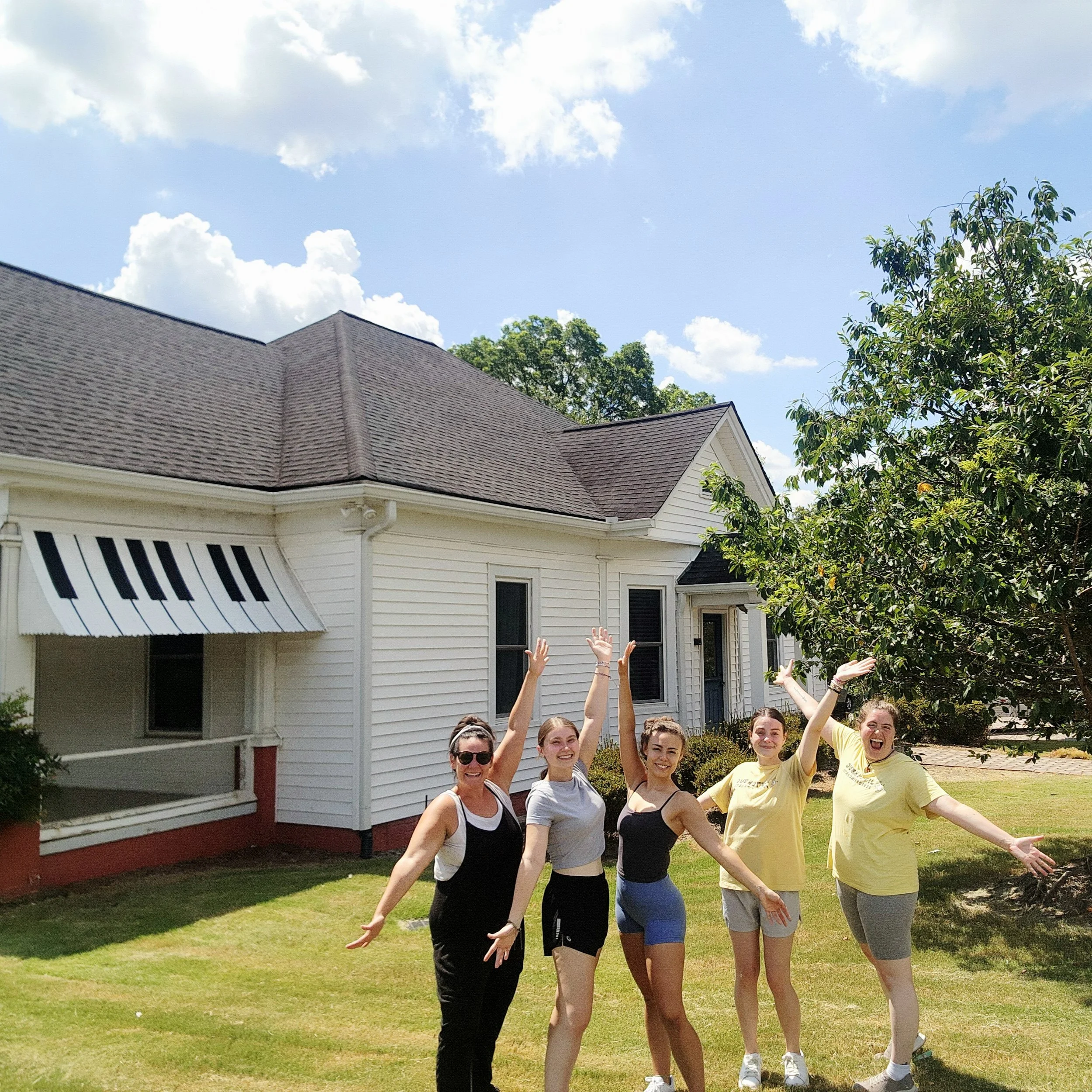 Five women standing outside in front of a white house with a brown roof, smiling, with arms raised in celebration, under a blue sky with white clouds.