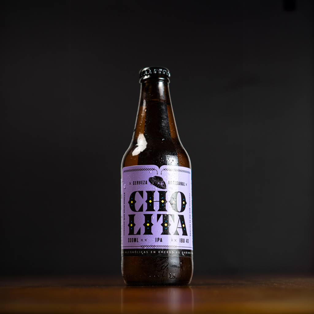 Botella de cerveza artesanal con etiqueta de color morado y letras grandes que dice 'Cholita' de estilo IPA en un fondo negro.