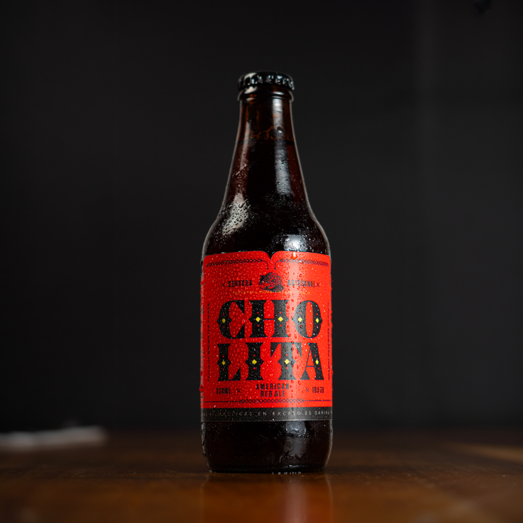 Botella de cerveza artesanal con etiqueta roja que dice Cholita American Red Ale en una superficie de madera, fondo negro.