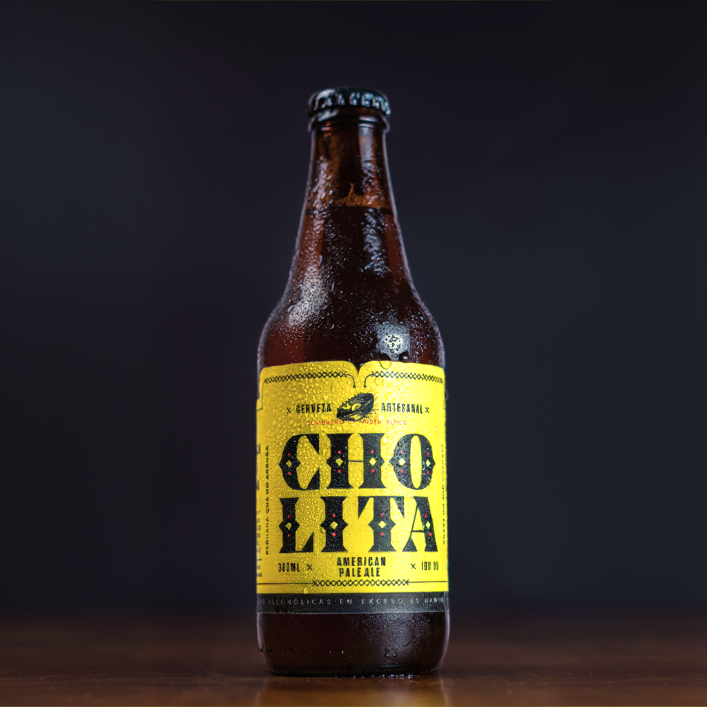 Botella de cerveza artesanal con etiqueta amarilla que dice 'Cholita' de estilo American pale ale en un fondo oscuro.