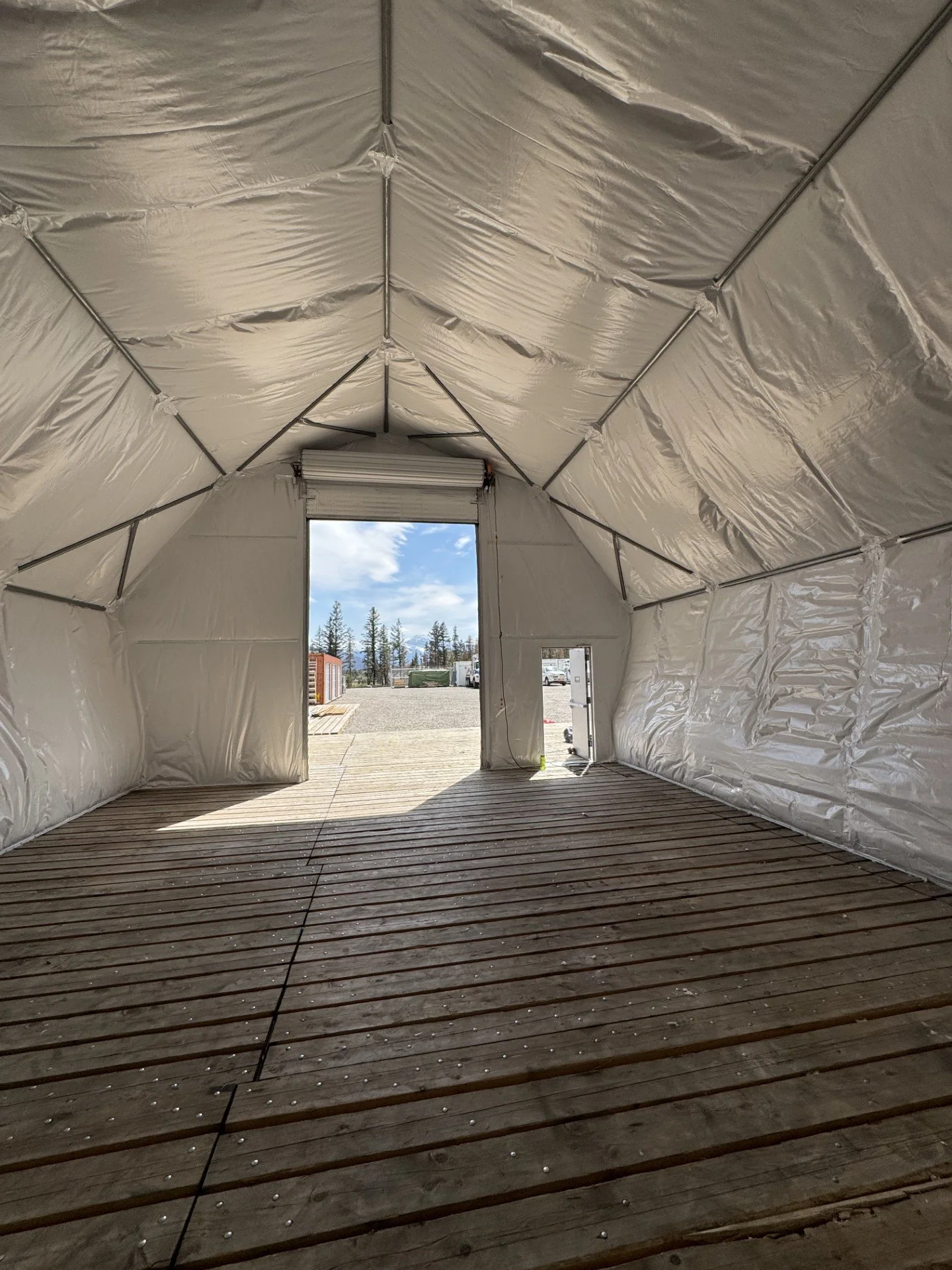 Interior of a temporary storage or event tent with a wooden floor, white insulated walls, and open doors showing an outdoor view with trees and blue sky.