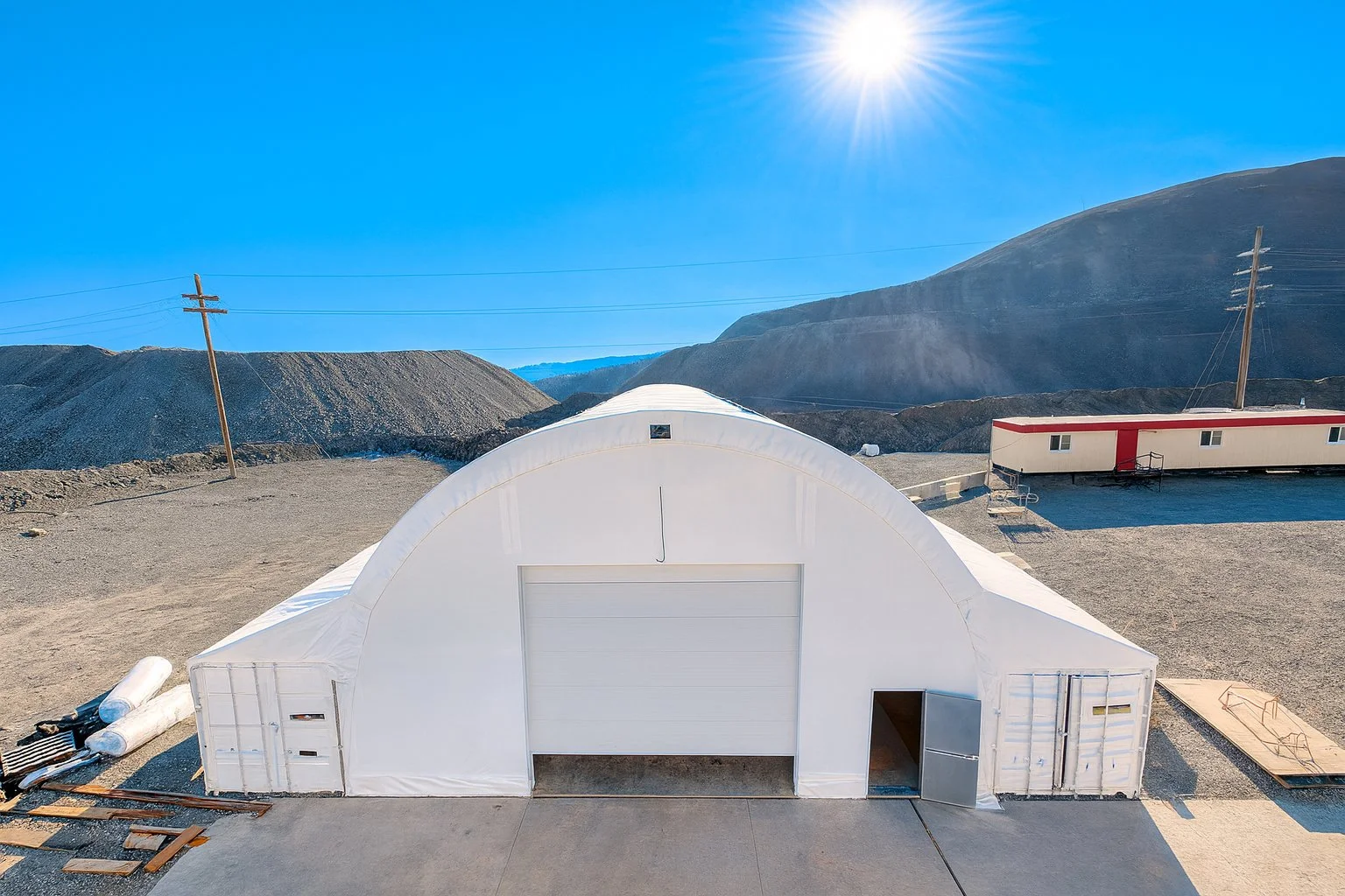 A large white storage tent outdoors in a desert-like area with mountains in the background, under a clear blue sky and bright sun.