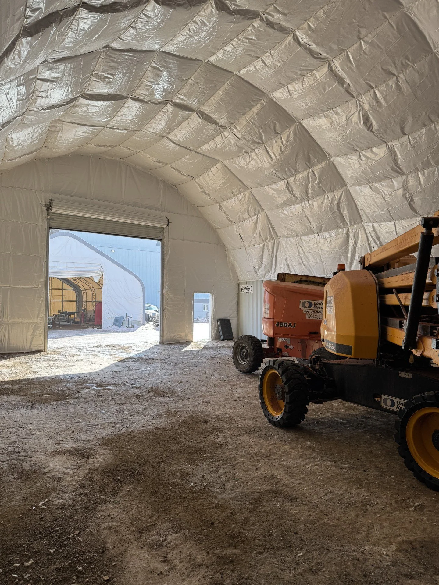 Inside a large white insulated tent with a dirt floor, construction equipment such as a Scissor lift is parked on the right side. In the background, there is a large open entrance, with another tent and some outdoor structures visible outside.