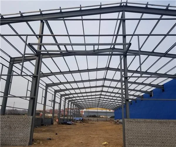 Steel framework of a building under construction with brick walls and a blue wall on the right side, on a dirt ground.
