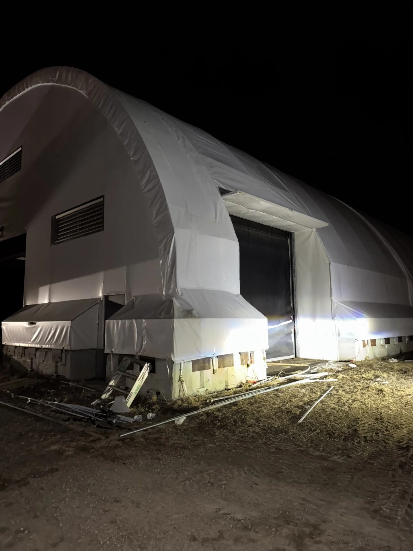 A large white inflatable structure under construction or maintenance at night with exposed ground and debris around it, illuminated by a bright light.