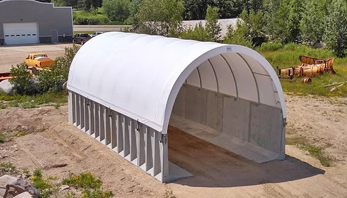 A large white fabric-covered storage shed with an open front, situated on a dirt lot with some green grass and bushes nearby, and construction or industrial equipment in the background.