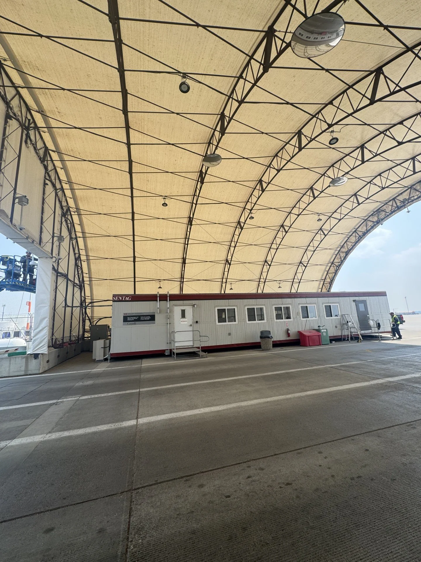 A large covered area with a curved fabric roof and exposed metal framework. There are temporary structures, including a white mobile office or container with windows and a door, on the ground. Some workers are visible in the background.