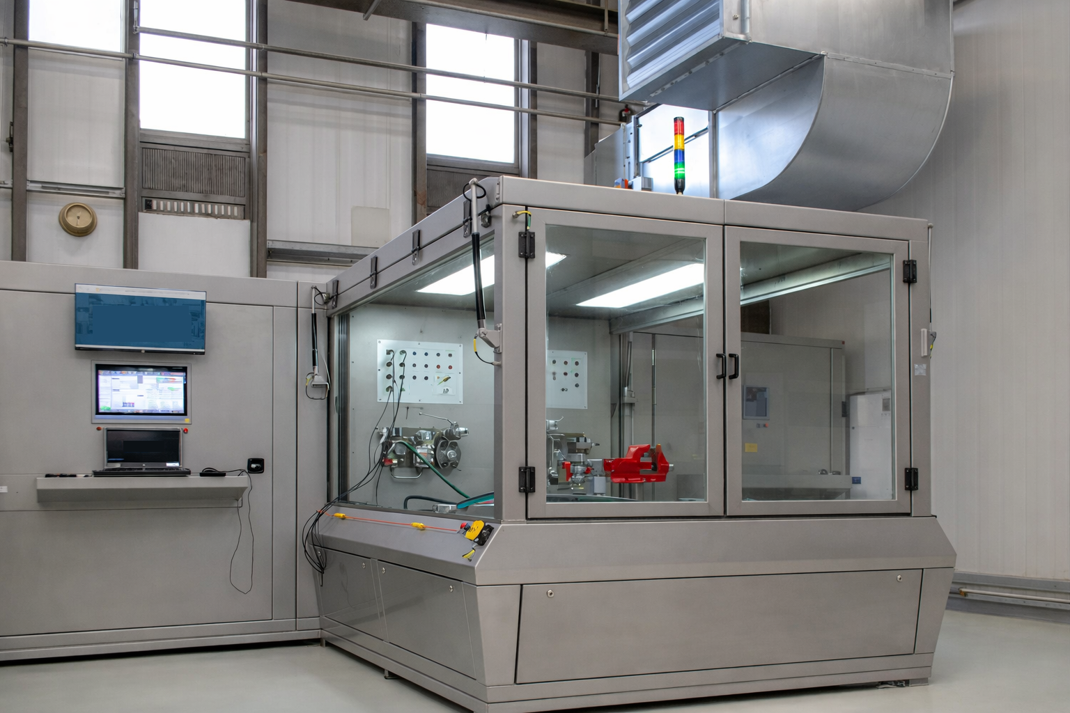 A large industrial machine with a transparent enclosed section in a spacious factory setting, with control screens and colorful indicator lights on top.