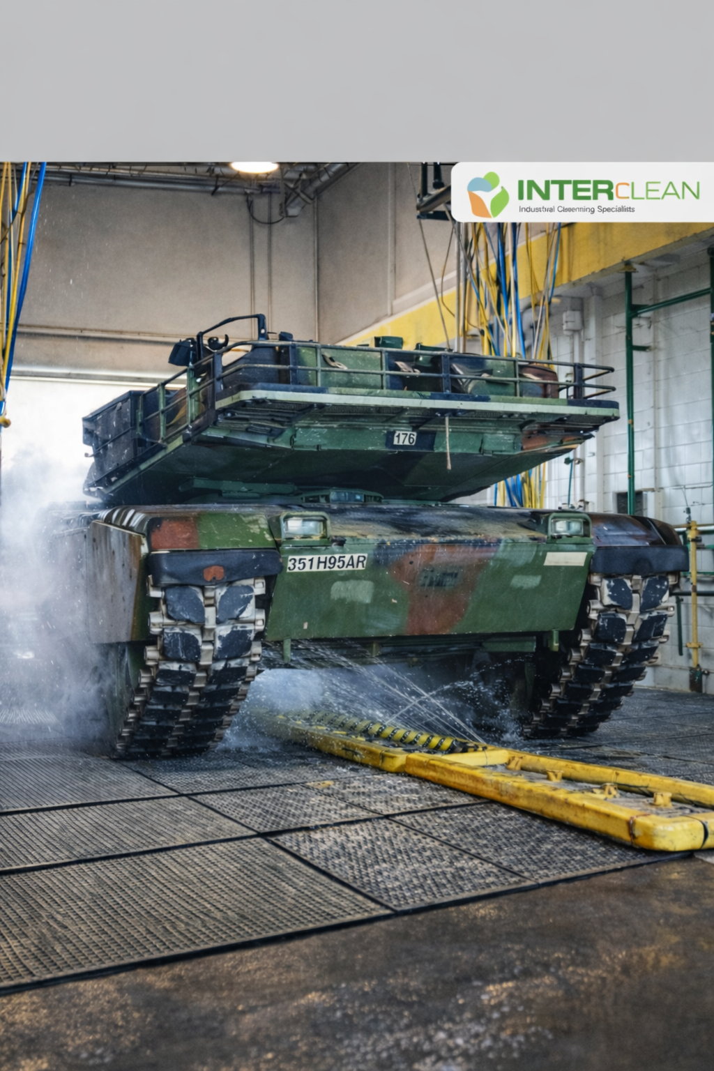 A military tank being pressure-washed inside a cleaning facility with yellow hoses hanging from the ceiling, and a sign that reads 'INTERCLEAN Industrial Cleaning Specialists.'