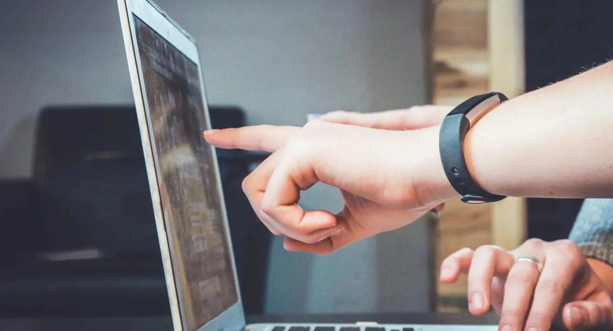 Close-up of a person's hand pointing at a laptop screen, with another person sitting nearby, indoors.