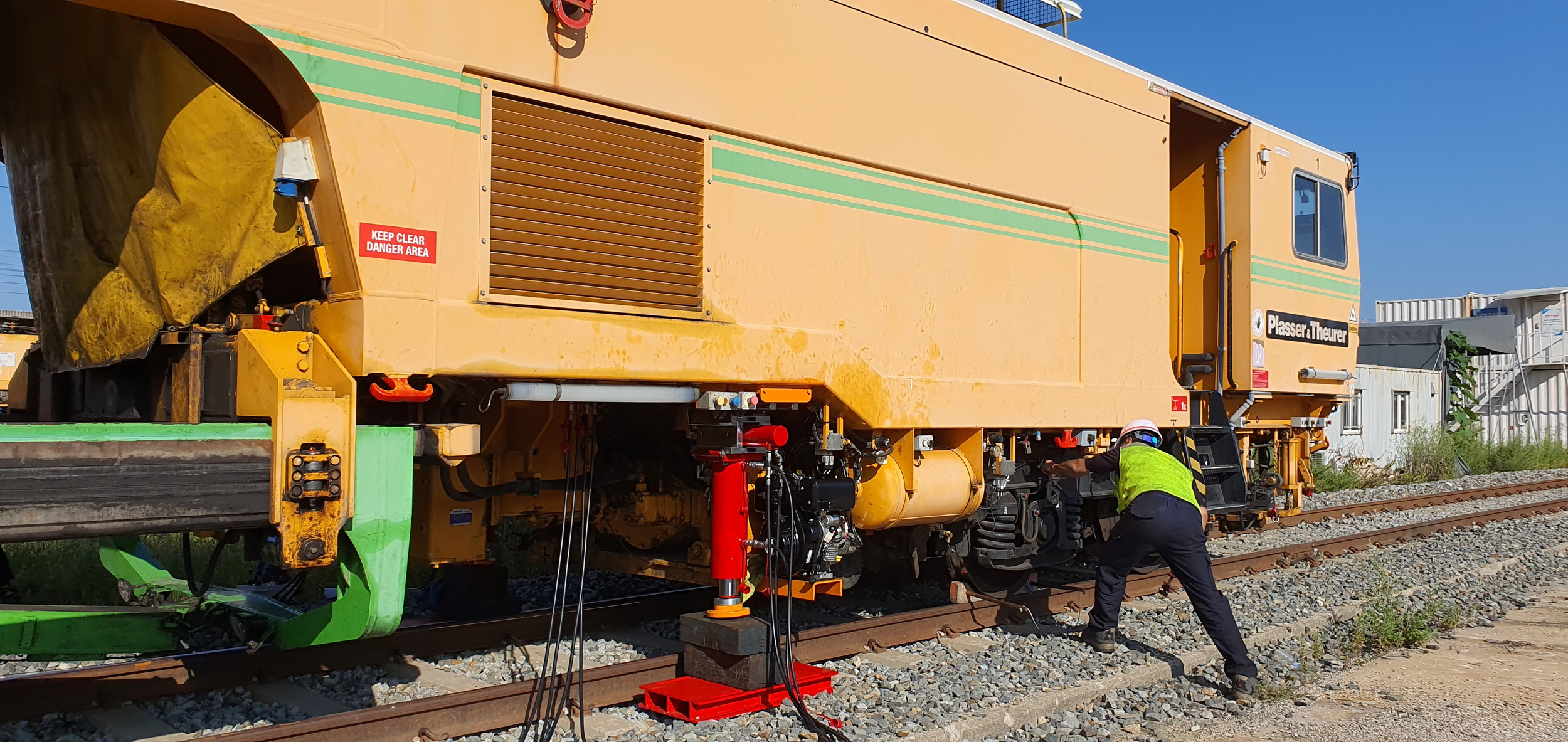 A railway maintenance worker in a reflective vest and helmet working under a yellow maintenance machine on train tracks in an outdoor area with clear blue skies and nearby structures.