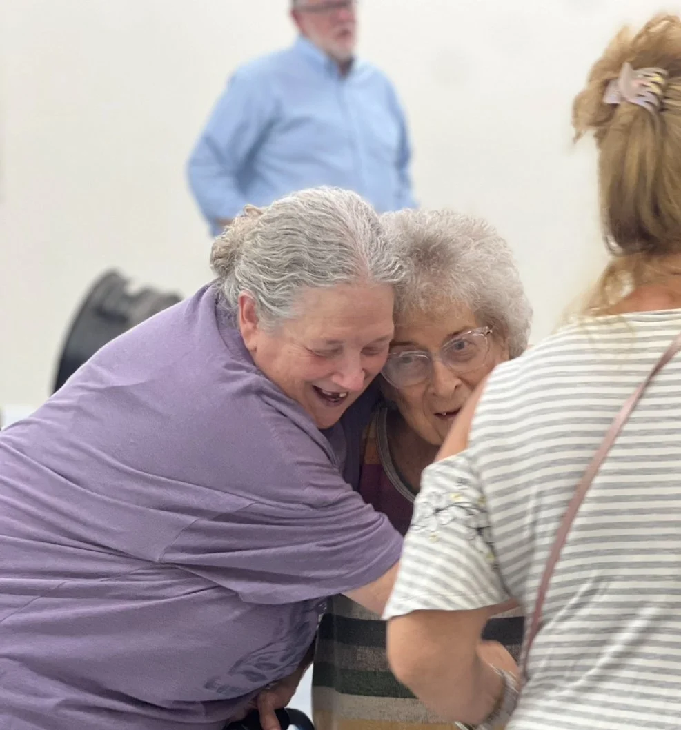 Three women are gathered together smiling and laughing, with two of them embracing each other, in a social setting. A man in a blue shirt stands in the background, slightly blurred.