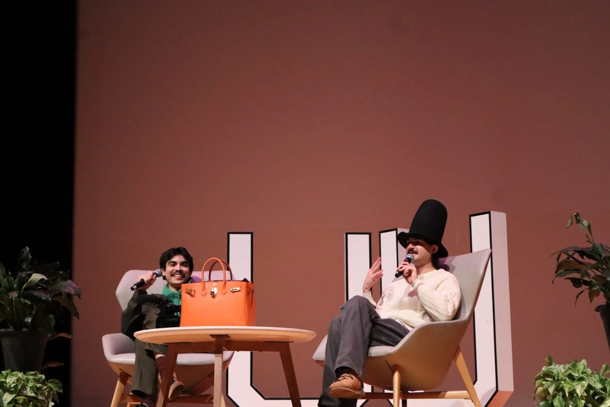 Two men sitting on stage in armchairs, having a conversation. The man on the left is smiling and holding a microphone, with an orange handbag on the table in front of him. The man on the right is wearing a tall black hat and a cream sweater, and is speaking into a microphone.