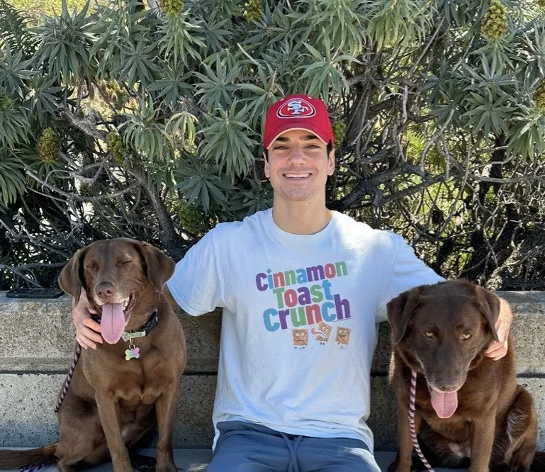 A young man sitting outdoors on a bench with two chocolate-colored dogs, all smiling with their tongues out, in front of a leafy green bush.