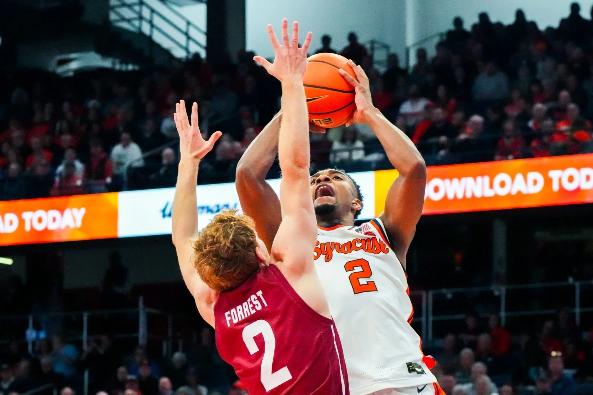 A basketball player from Syracuse University, wearing jersey number 2, attempting a shot while being guarded by a player from Colgate University during a game with a large crowd in the background.