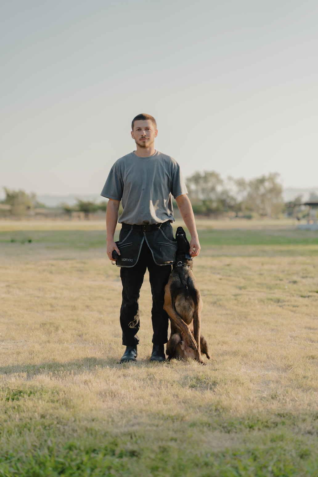 A young man standing outdoors on a grassy field holding a German Shepherd puppy on a walk, with sparse trees and a clear sky in the background during daytime.