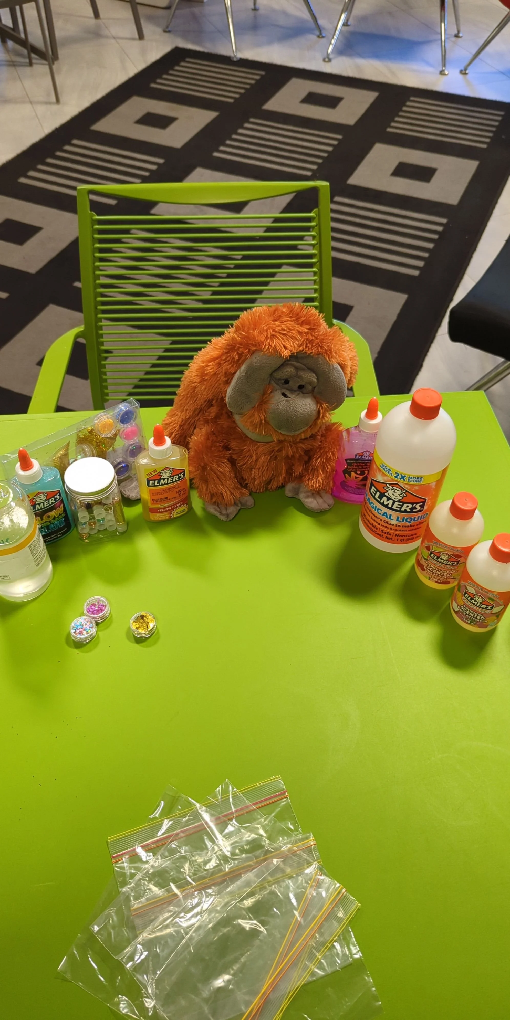 Table with craft supplies including bottles of glue, small containers of glitter, and start of small plastic bags. A plush orangutan toy is sitting on the table. In the background, there is a patterned black and white rug and green and black chairs.