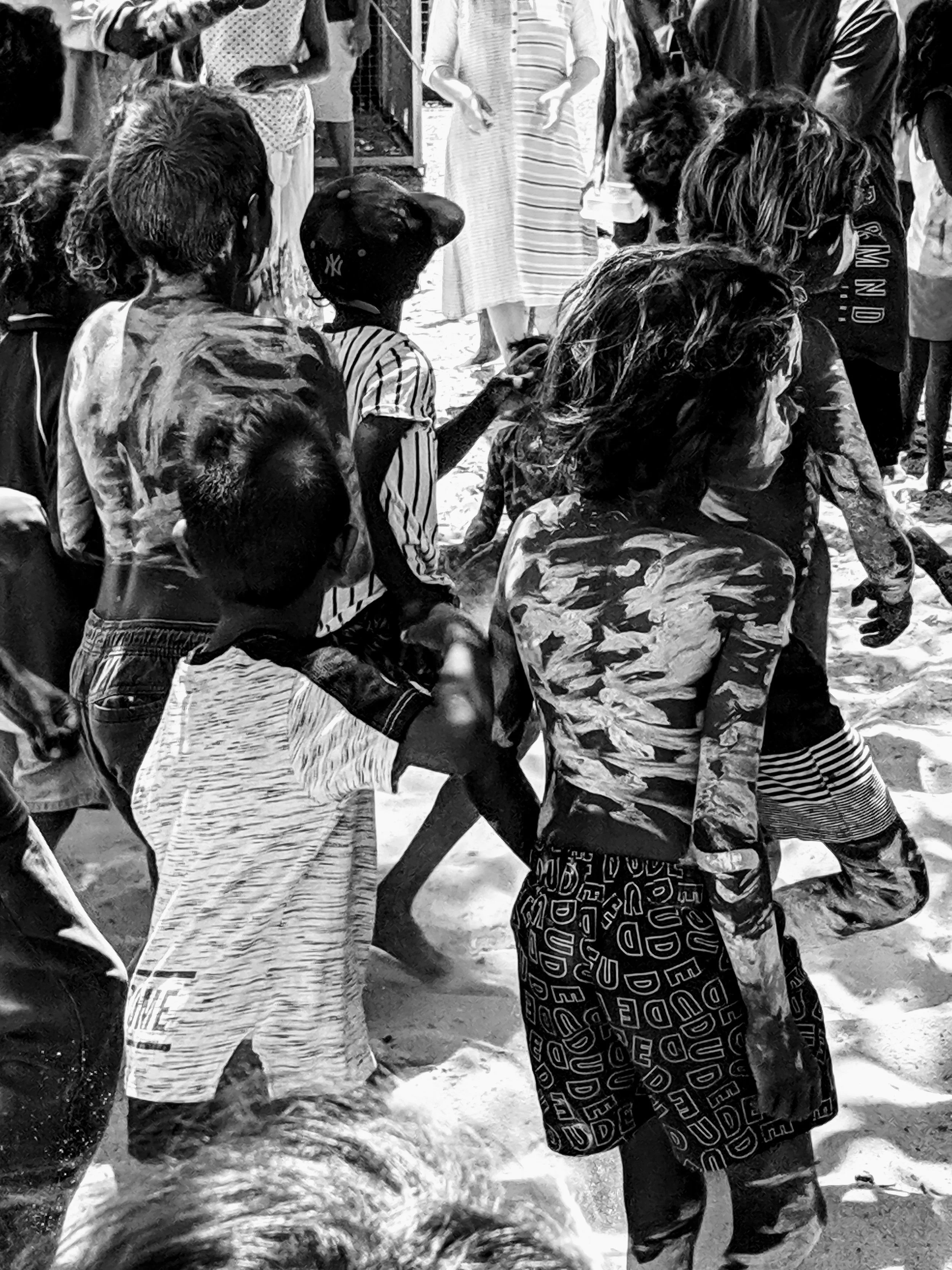 Children and adults gather outdoors, some holding hands, on sandy ground, engaging in a group activity.