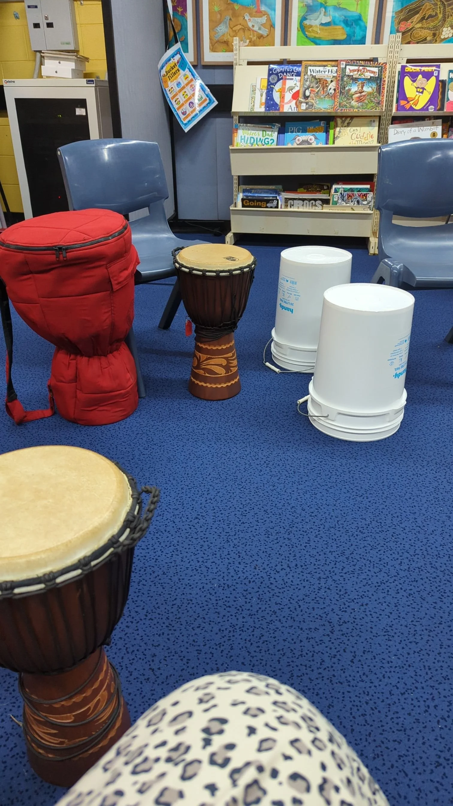 A cozy reading area in a library with blue carpet, two chairs, a red backpack on a chair, two djembe drums, buckets, and a bookshelf filled with children's books. Artworks are displayed on the wall in the background.