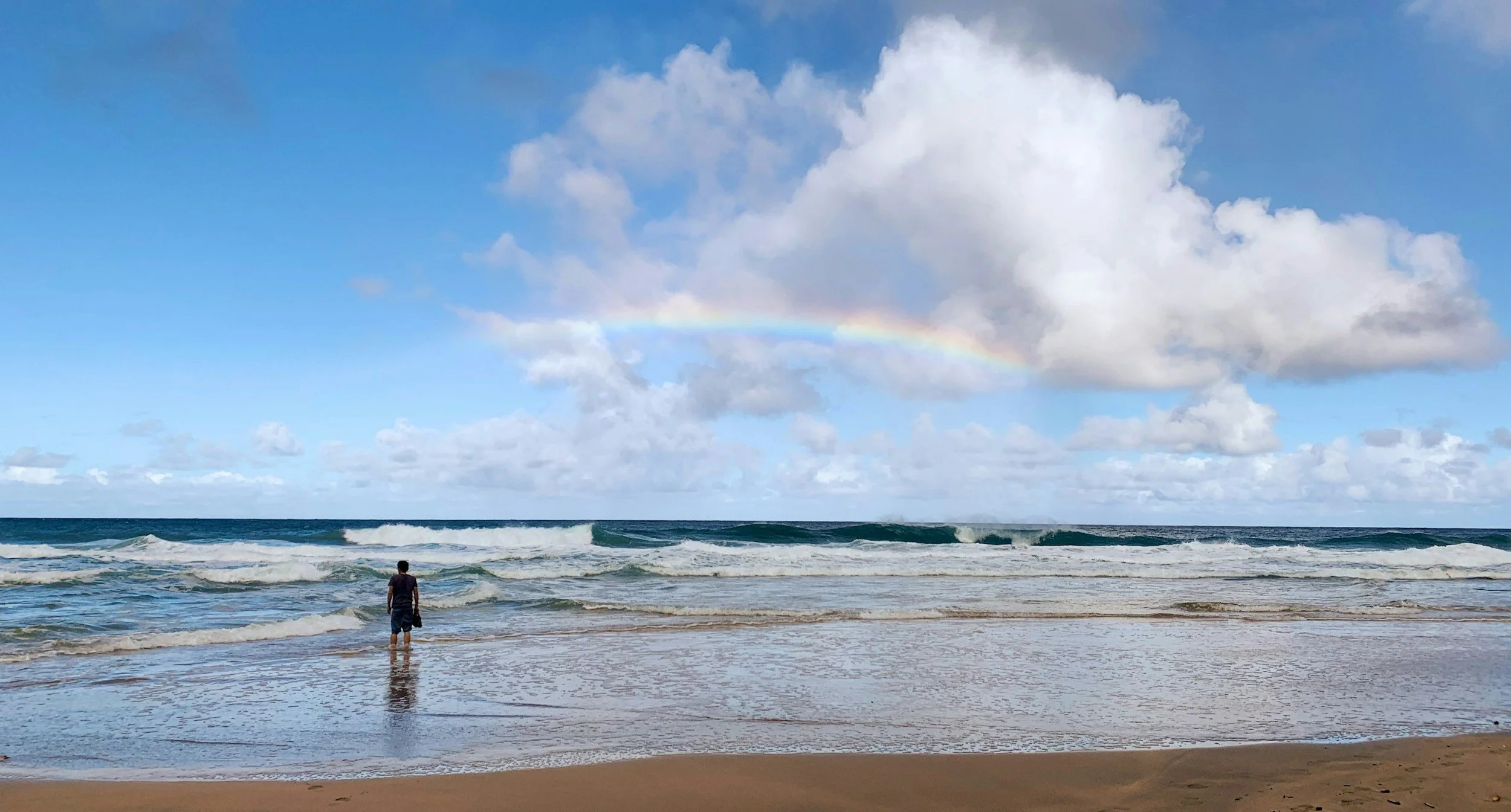 A person standing at the shoreline of a beach, facing the ocean under a sky with clouds and a faint rainbow.