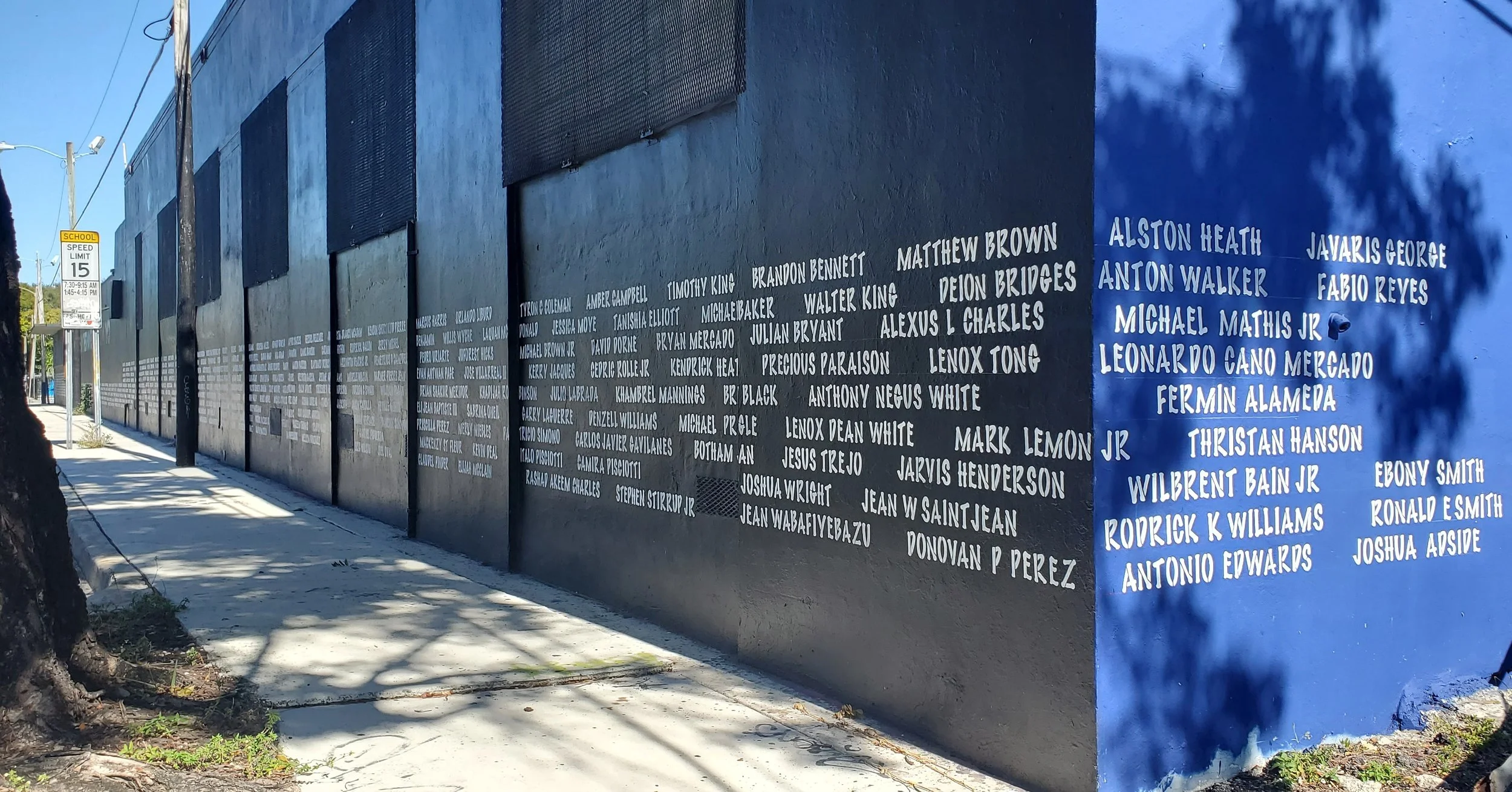 Image depicts a long exterior wall painted black and blue, it displays dozens of names painted in white lettering. The names are arranged in rows across the side of the building, resembling a memorial.