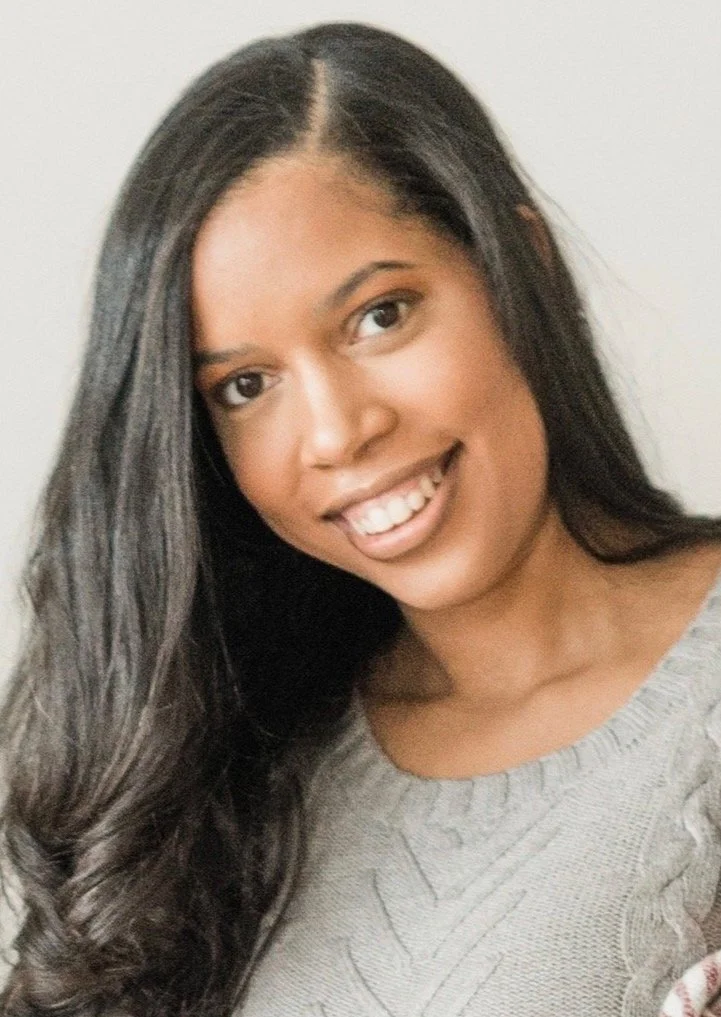 Close-up of a woman with long dark hair smiling, wearing a gray sweater, against a plain background.