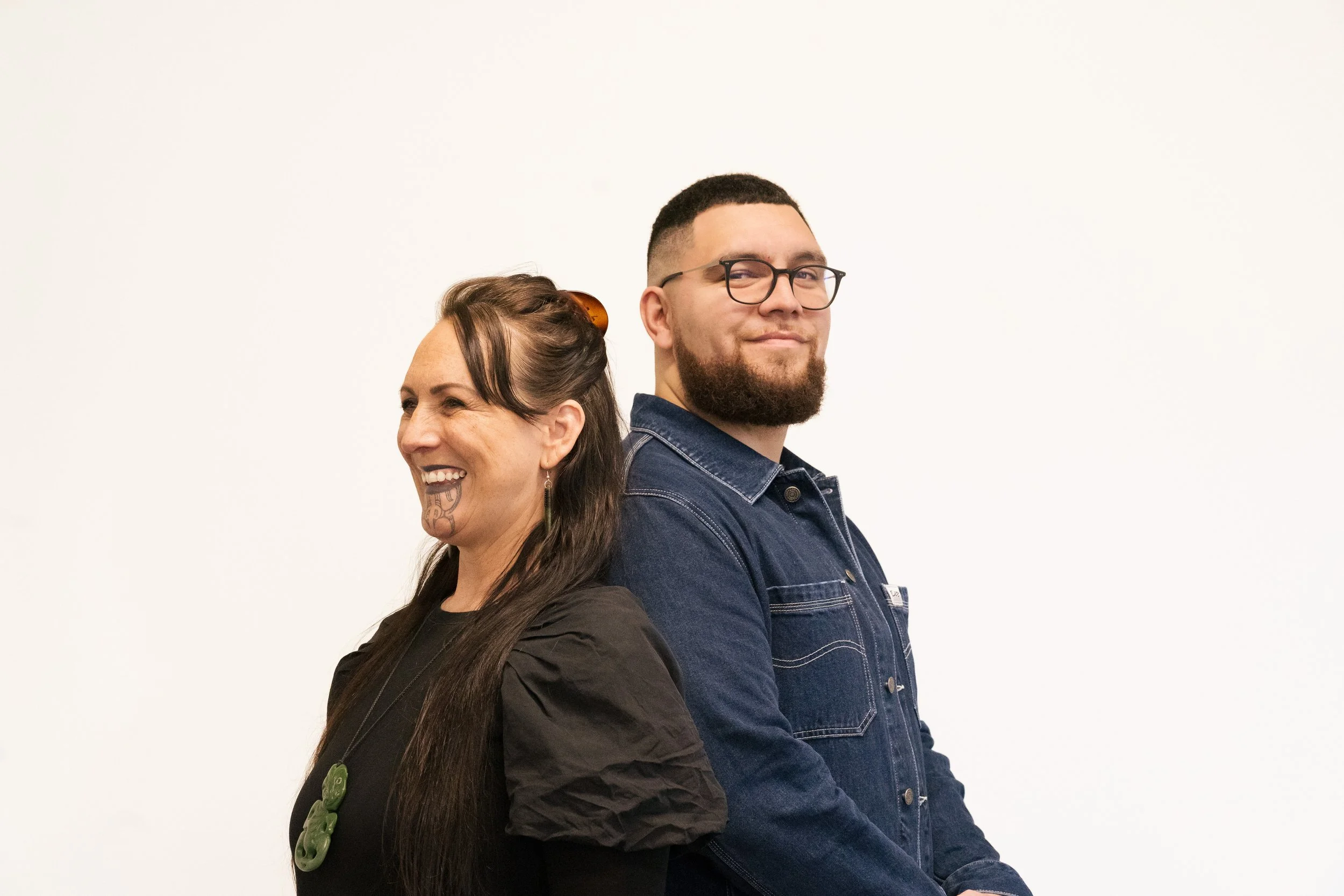 Jacob (Director) and Nicky (Pou Kōrero) are standing in front of a blank background, back to back. Nicky is looking off to the side and Jacob is looking at the camera.