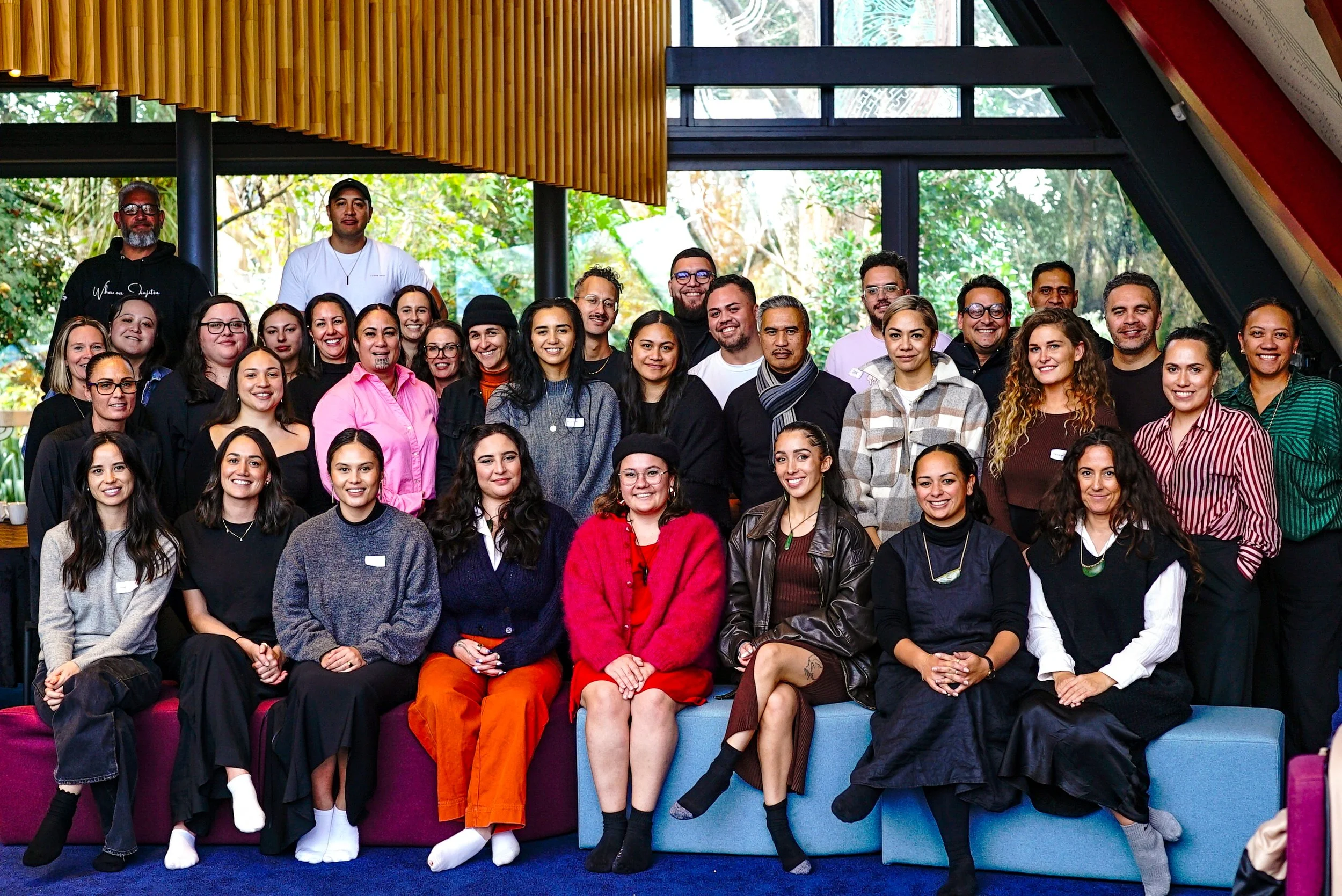Diverse group of Māori gathered indoors for a group photo, with some seated in front and others standing behind. The background features large windows showing greenery outside, and the room has an interior design with wooden and sloped roof elements.