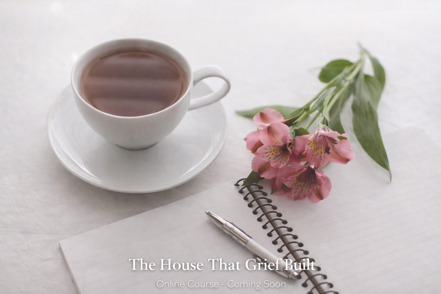 A white cup filled with tea on a saucer next to a pink flower with green leaves and a silver pen on a spiral notebook with lined paper, all on a white surface.