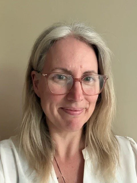 A woman with long blonde hair, glasses, and a light-colored shirt smiling in front of a plain beige wall.