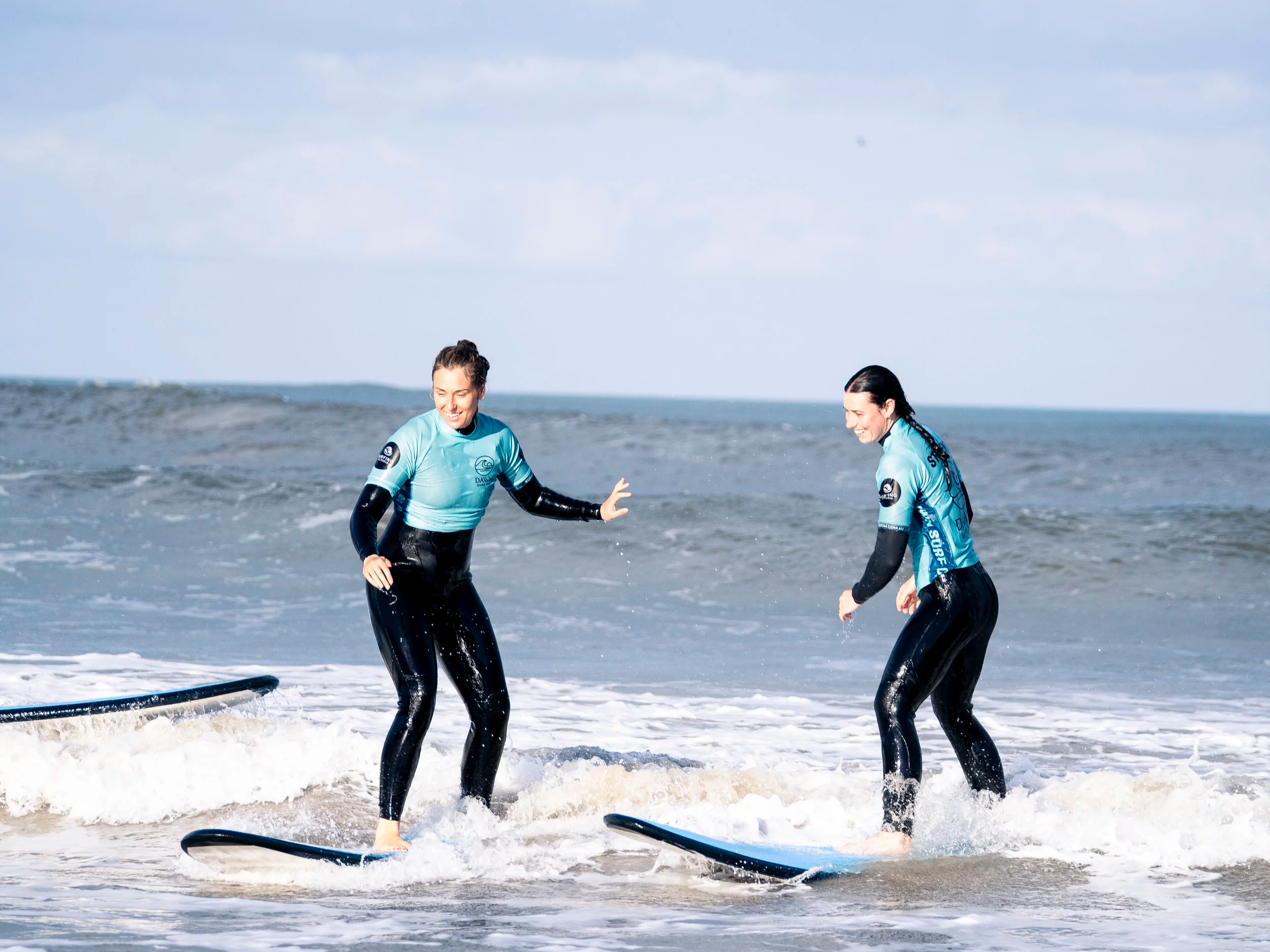 Two women in wetsuits surfing on the beach, smiling and having fun in the ocean waves.