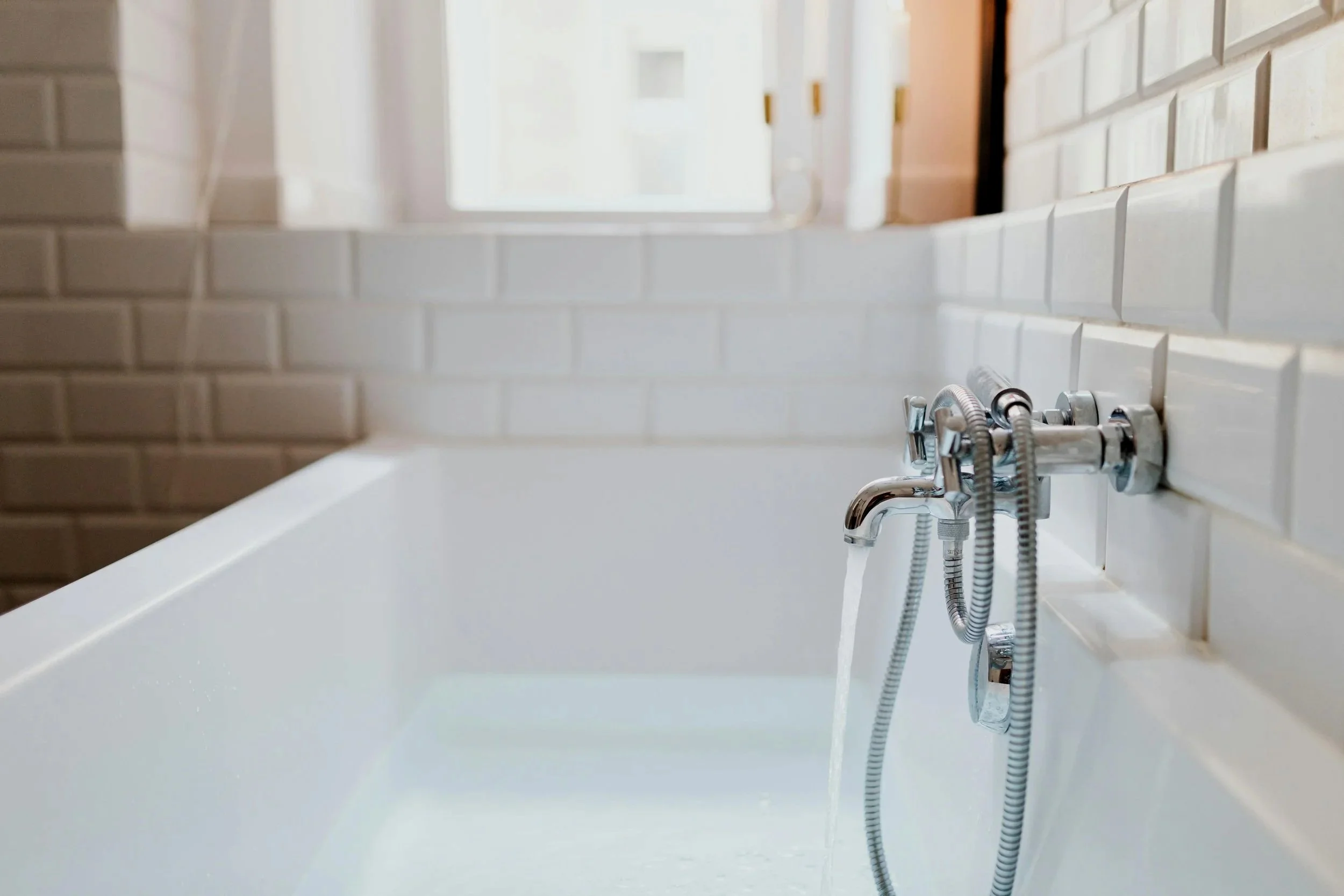 Close-up of a modern bathtub faucet with water flowing into a white bathtub, surrounded by white tiled walls in a bathroom with a window in the background.