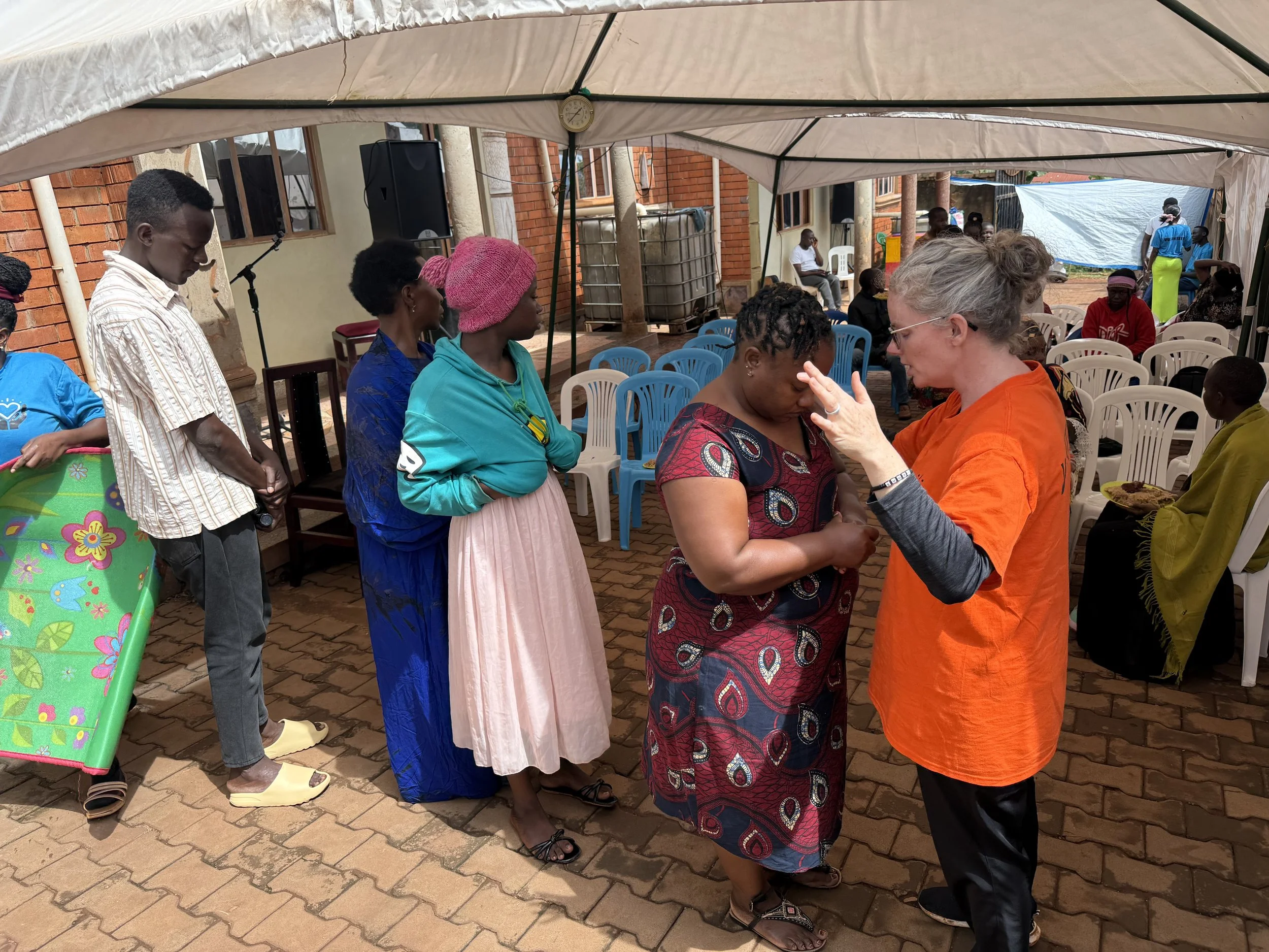 A group of people standing in a line under a tent during an outdoor event. A woman with gray curly hair and glasses is touching the forehead of a woman in patterned clothing. Others wait with heads bowed or eyes closed, and some are seated in white plastic chairs. The setting is a brick-paved area with buildings and other tents in the background.