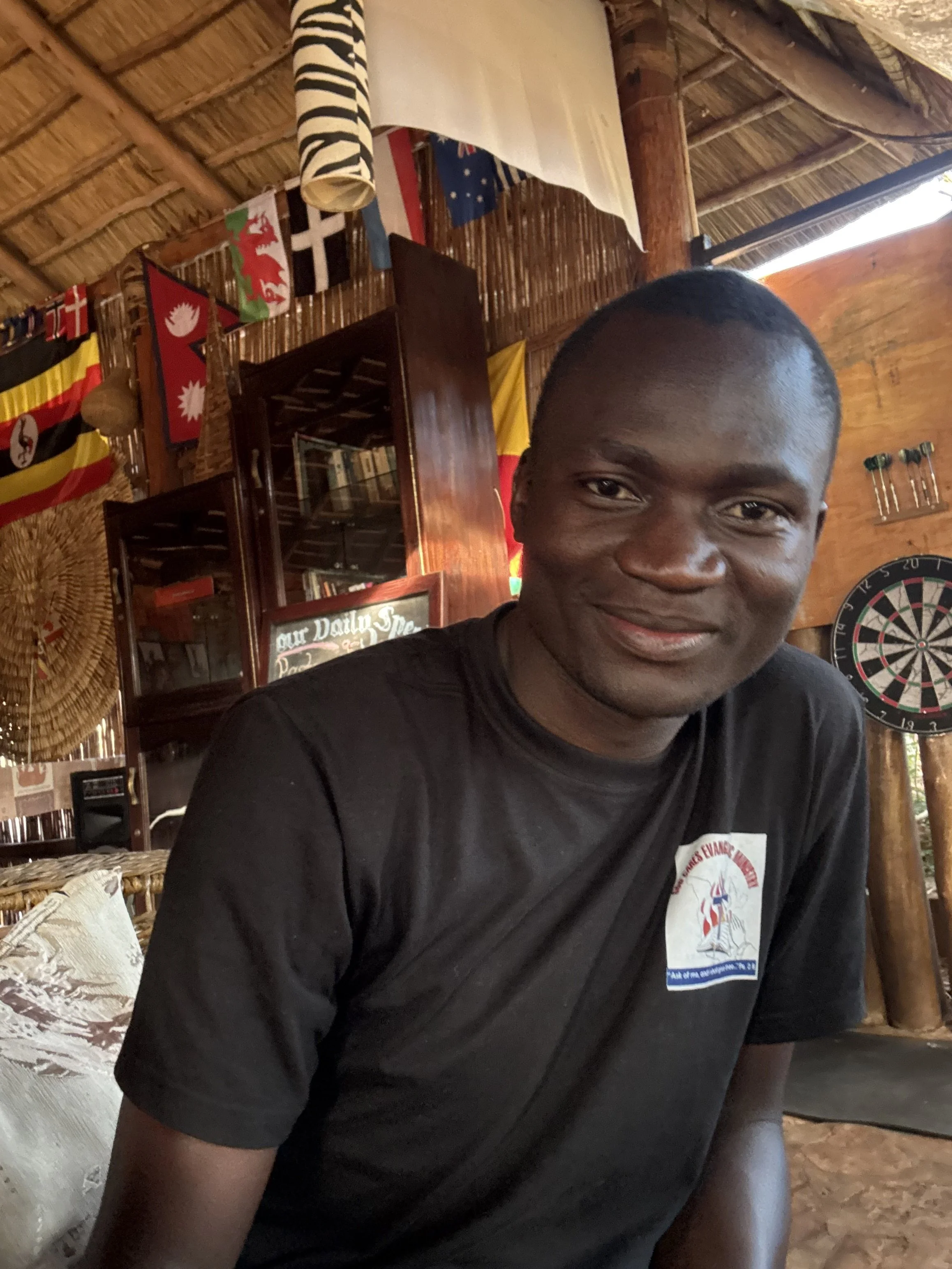 A young man with dark skin and a shaved head, smiling, sitting in a rustic wooden room with flags and dartboards on the walls.