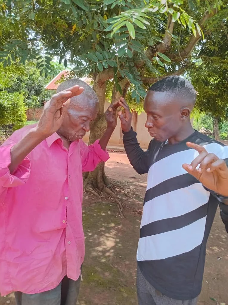 Two men with closed eyes and hands raised, appearing to be praying or engaging in a spiritual activity outdoors under a large leafy tree.